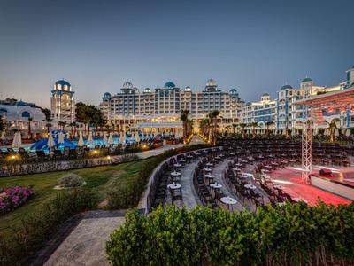 Grand complexe hôtelier éclairé avec scène en plein air et sièges extérieurs au crépuscule.