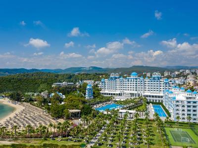 Großes Strandhotel mit Pools, Tennisplätzen und grünen Gartenanlagen vor blauem Himmel.