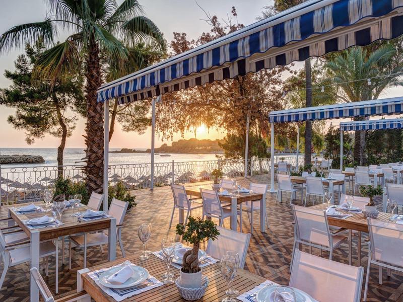 Terrasse de restaurant en bord de mer avec tables sous des auvents au coucher du soleil.