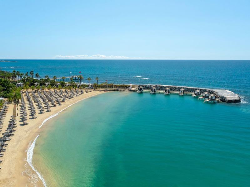 Plage courbée avec chaises longues et parasols, jetée s'étendant dans une mer turquoise.