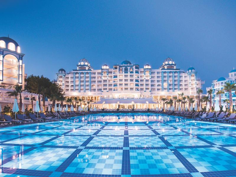 Piscine illuminée devant un grand hôtel blanc sous un ciel bleu au crépuscule.