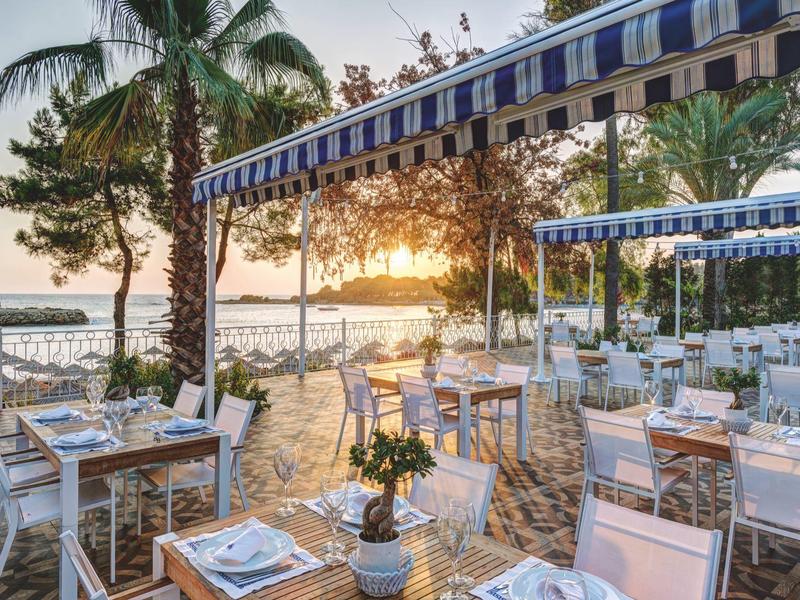 Terrasse de restaurant en bord de mer au coucher du soleil, tables blanches et parasols bleus.