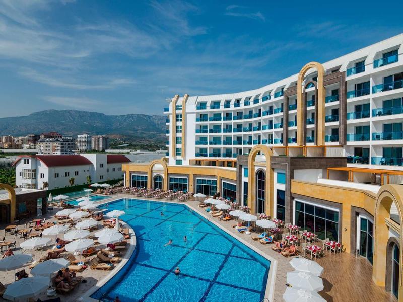 Moderní hotel s velkým venkovním bazénem a sluneční terasou pod modrou oblohou.