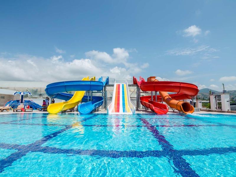 Barevný bazén se třemi vodními skluzavkami a čistou modrou vodou pod částečně zataženou oblohou.