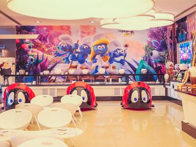 Buntes Café mit Smurfs-Wandbild und roten Smurfs-Kindersitzen, weiße Tische und Stühle.
