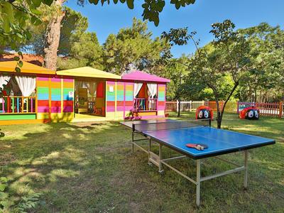Bunter Holzbungalow mit rotem Dach auf grüner Wiese, davor Tischtennisplatte und umgeben von Bäumen.
