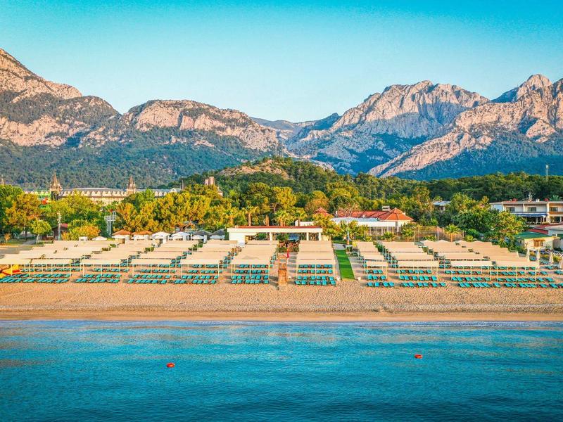Hotel de playa con sombrillas y tumbonas frente a montañas y cielo claro junto al mar.