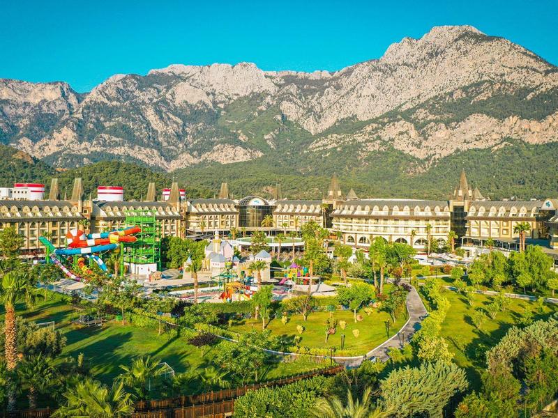Complejo hotelero frente a una cadena montañosa con jardines verdes y palmeras bajo un cielo despejado.