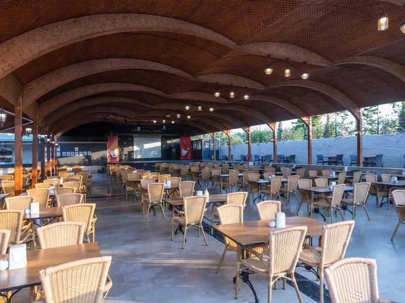 Velká krytá venkovní plocha s mnoha stoly a židlemi pod dřevěnou obloukovou konstrukcí.