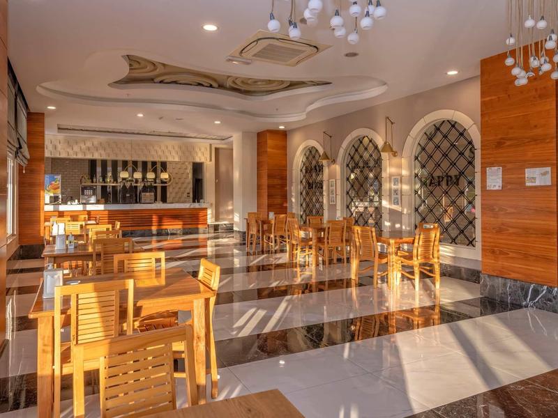 Světlá hotelová restaurace s dřevěným nábytkem, mramorovou podlahou a velkým barem v pozadí.