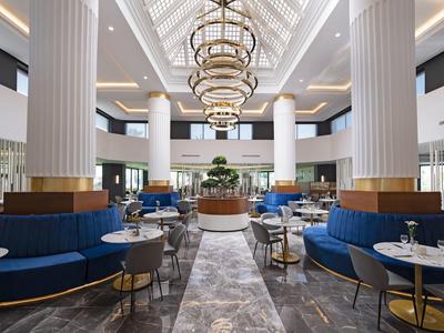 Großer, moderner Lounge-Bereich mit blauen Sofas, runden Tischen, weißen Säulen und Kronleuchtern.