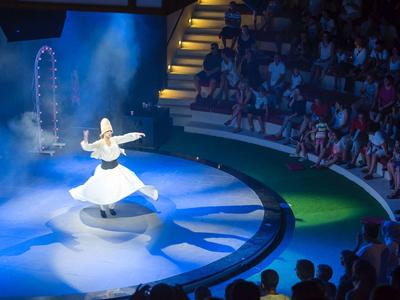 Eine drehende Bauchtänzerin im weißen Kostüm auf einer Bühne vor Publikum im Theater.
