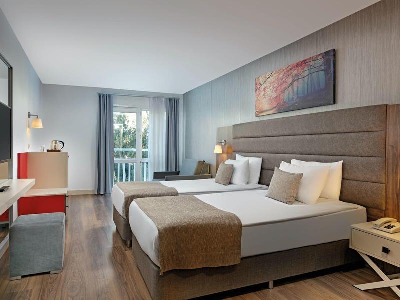 Moderní hotelový pokoj se dvěma jednolůžky, dřevěnou podlahou, psacím stolem a balkónovými dveřmi se závěsy.