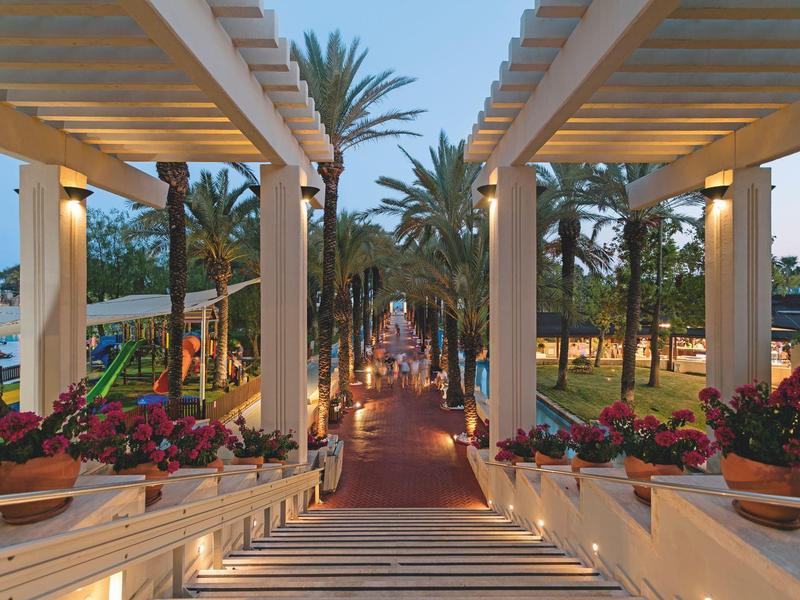 Cesta se schody, pergolami a květináči vedoucí do zahrady s palmami za soumraku.