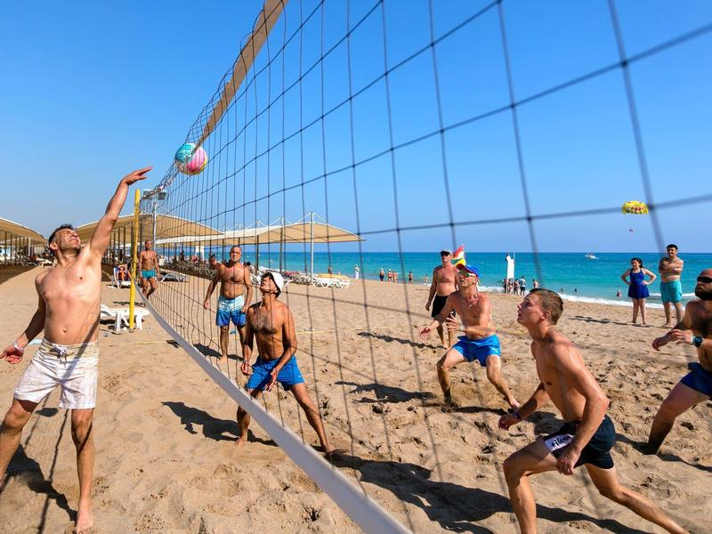 Skupina mužů hrající plážový volejbal na slunečné písečné pláži u průzračného modrého moře.