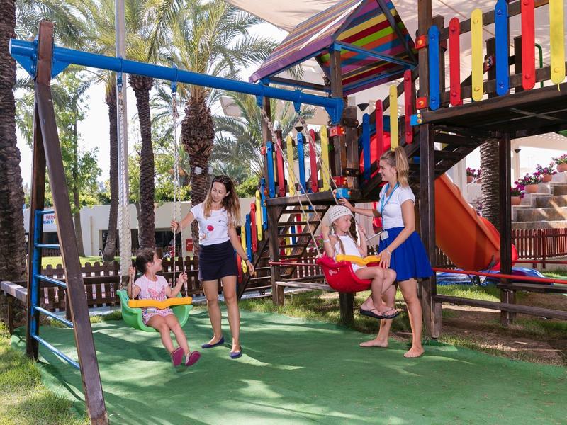 Děti a dospělí si hrají na barevných houpačkách na slunném venkovním prostranství s palmami.