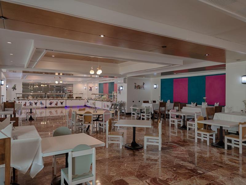 Prostorná hotelová jídelna s bílými stoly a židlemi, barevnými nástěnnými panely a dlažbou na podlaze.