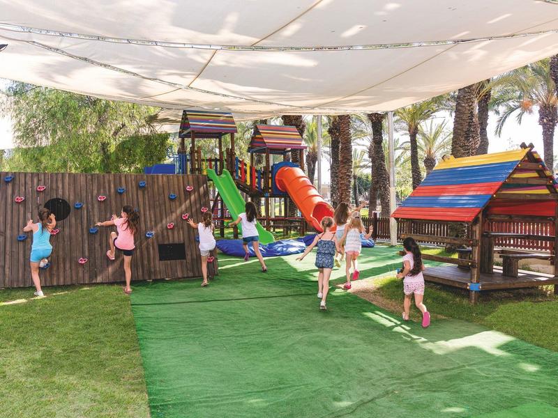 Děti si hrají ve stíněném venkovním hřišti s klouzačkami a houpačkami na zelené trávě.