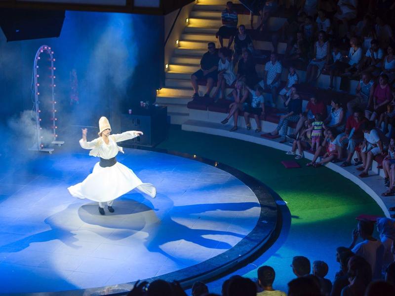 Tanečník v bílém točí se na kulaté plošině s modrým osvětlením a sedícím publikem kolem.