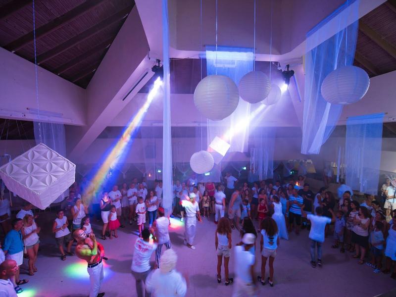 Přecpaná párty v interiéru s barevnými světly a zavěšenými kulatými ozdobami.