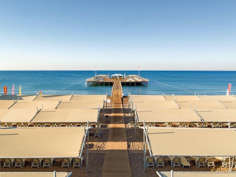 Vista de un largo muelle y tumbonas sombreadas en la playa con el mar azul al fondo.