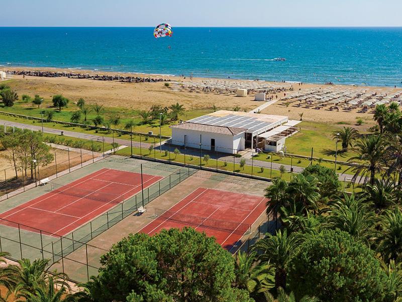 Vista de canchas de tenis, palmeras y playa con mar azul al fondo bajo un cielo soleado.