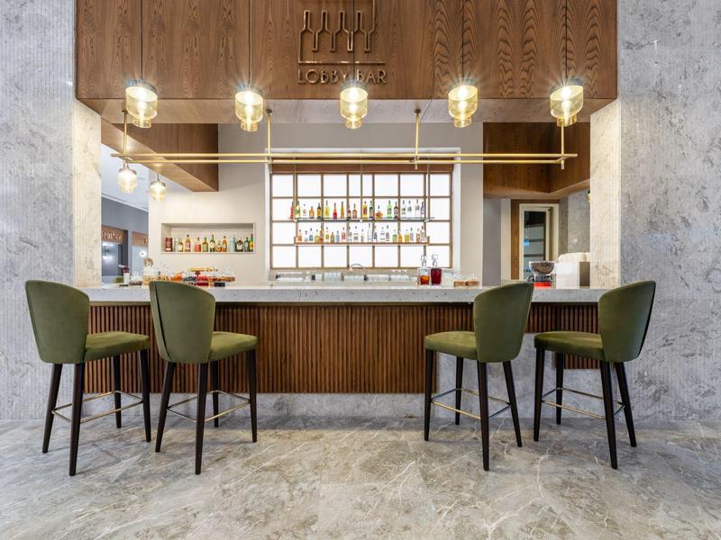 Área moderna de bar de hotel con sillas acolchonadas verdes y mostrador de madera bajo luz cálida.