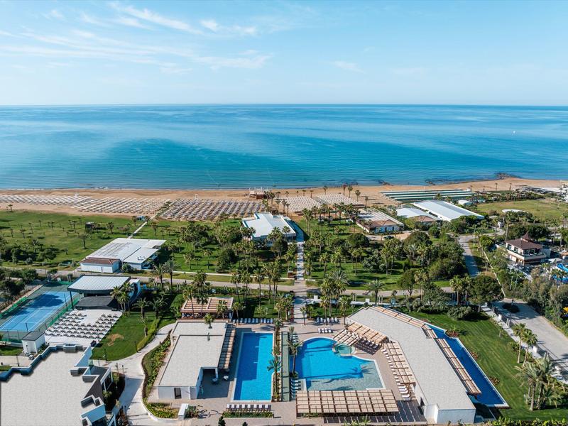 Vista aérea de un complejo hotelero con varias piscinas cerca de una larga playa de arena y mar azul.