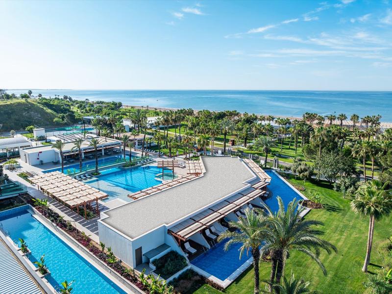 Complejo hotelero moderno con piscina, rodeado de palmeras y vista al mar en un día soleado.