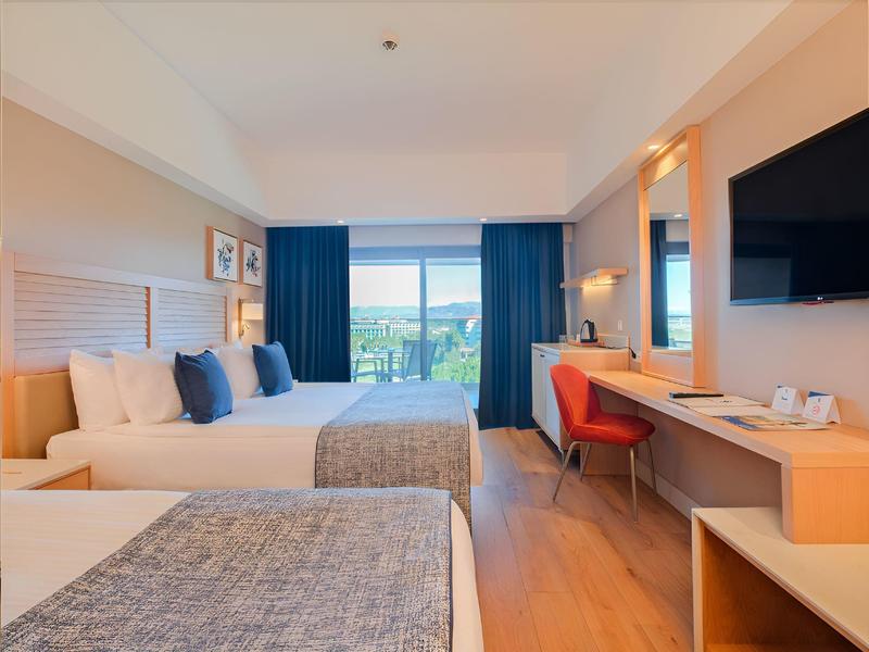 Habitación de hotel luminosa con dos camas, escritorio, silla y gran ventana con vista a la naturaleza.