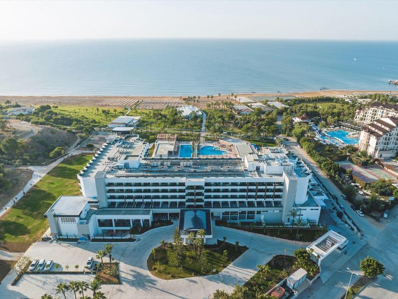 Gran hotel con piscina cerca de la playa, rodeado de árboles y estacionamiento, mar al fondo.