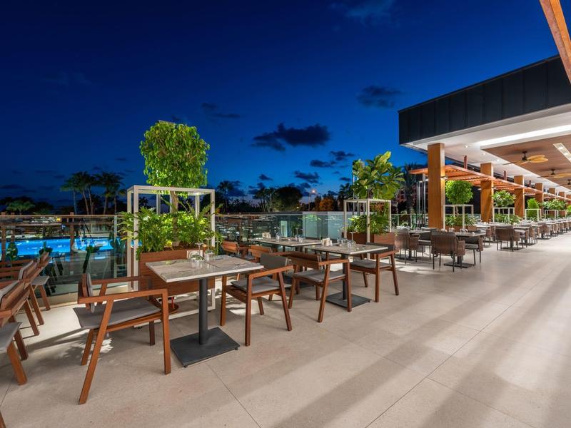 Restaurante al aire libre en el hotel con sillas y mesas de madera junto a una piscina iluminada por la noche.
