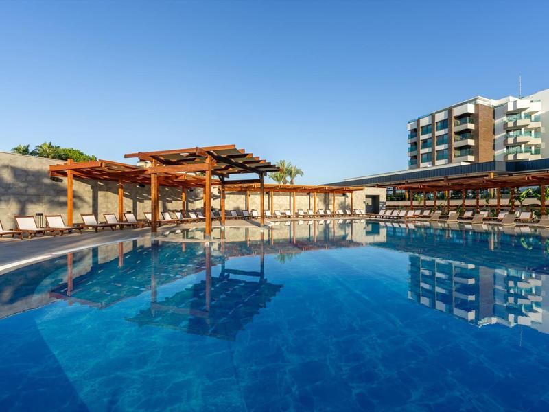 Gran piscina azul de hotel con tumbonas y edificios modernos al fondo bajo cielo despejado.