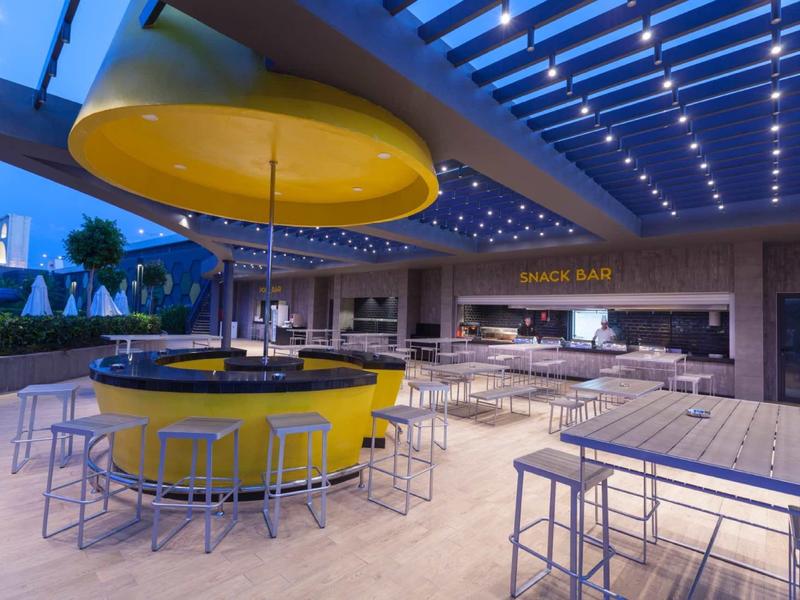 Modern hotel bar with round yellow counter, stools, and covered outdoor area at dusk.