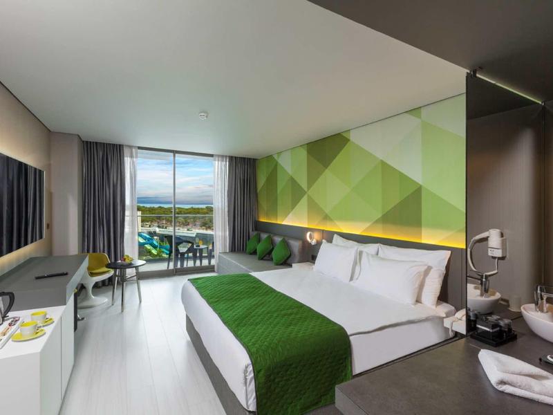 Modern hotel room with large bed, green accents, and balcony overlooking the sea