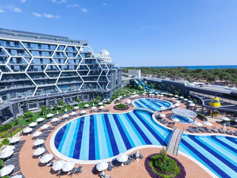 Large hotel with unique facade, round pools, and many loungers under a clear sky.