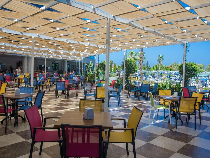 Outdoor restaurant with colorful chairs under a large canopy and palm trees in view.