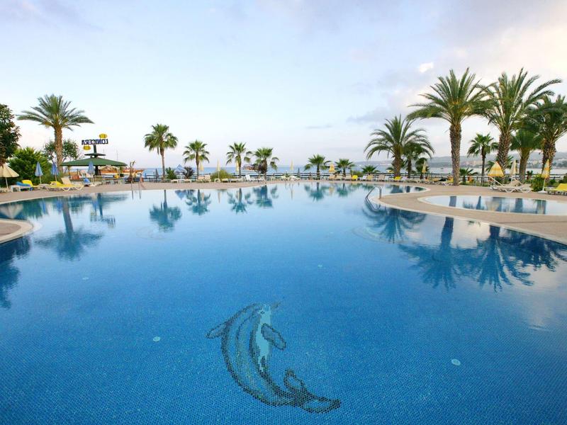 Large swimming pool with dolphin mosaic, surrounded by palm trees and lounge chairs under a blue sky.