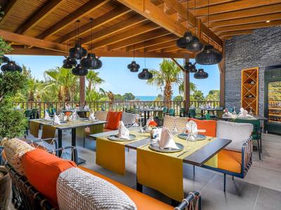 Open eetgedeelte met gedekte tafels, kleurrijke stoelen en uitzicht op tropische planten en zee.