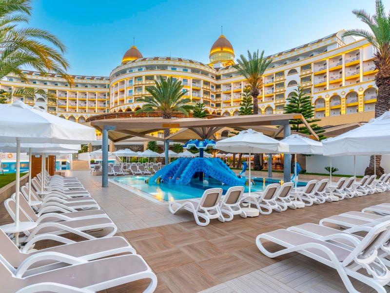 Groot resort met zwembad, palmbomen en veel witte ligstoelen onder een heldere hemel.