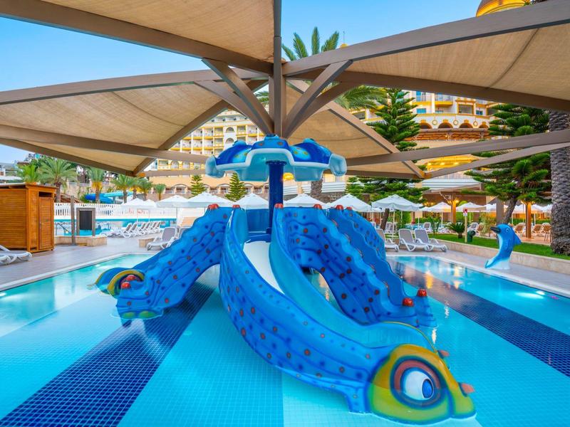 Kleurrijke kinderspeelplek met glijbaan in een hotelzwembad onder parasols.