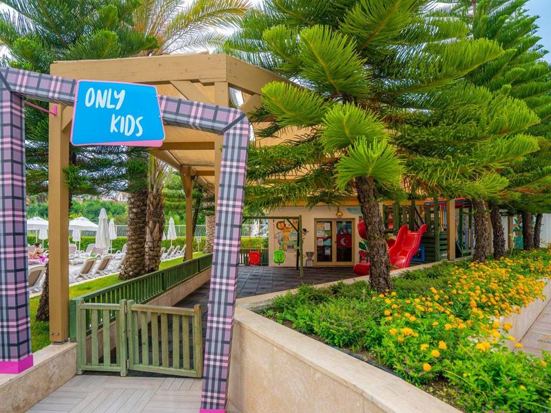 Ingang naar een speelruimte voor kinderen met een blauw bordje 'Only Kids', omgeven door bomen en bloemen.