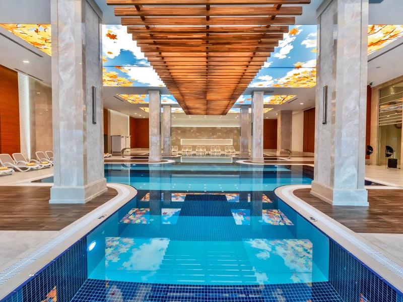 Moderne overdekte zwembadruimte met blauw water, witte zuilen en houten plafondpanelen.