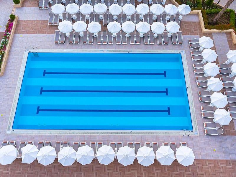 Blauw rechthoekig zwembad omgeven door witte parasols en terrasmeubilair.