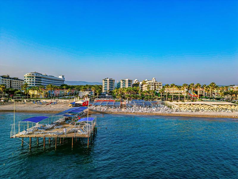 Een lange pier loopt over kalm water naar een zandstrand met hotels en palmbomen op de achtergrond.