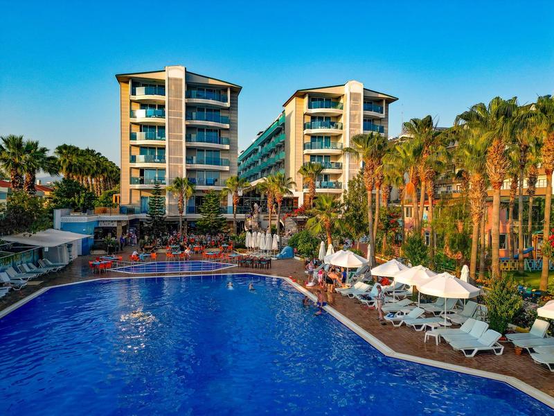 Modern hotelcomplex met groot zwembad, ligstoelen en palmbomen onder heldere lucht.