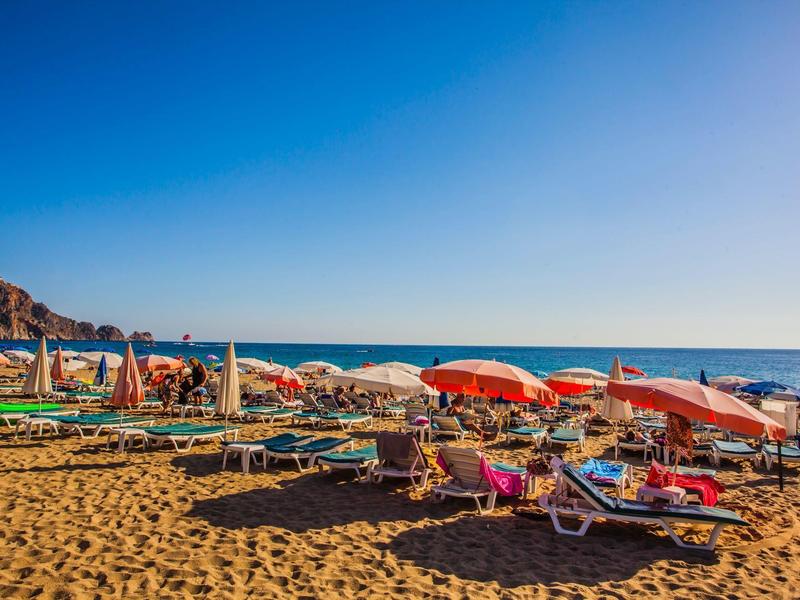 Sandstrand mit Liegen und roten Sonnenschirmen, blauer Himmel und Meer im Hintergrund.