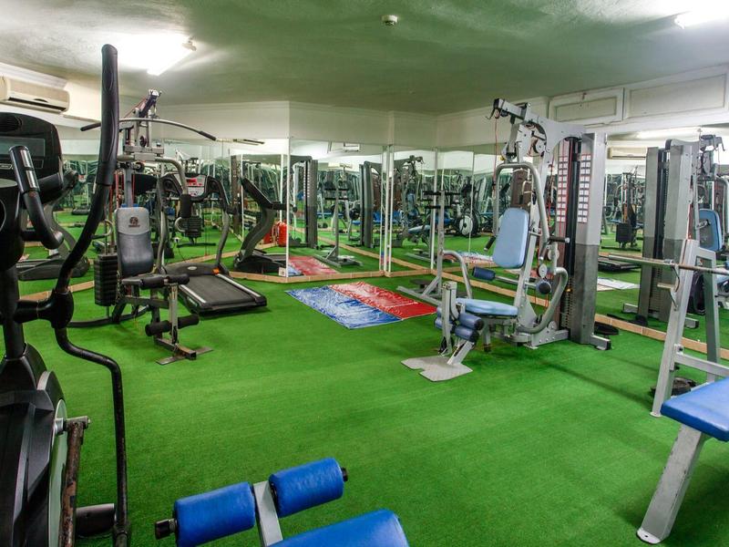 Fitnessraum mit grüner Teppichfläche, verschiedenen Trainingsgeräten und großen Spiegeln an der Wand.