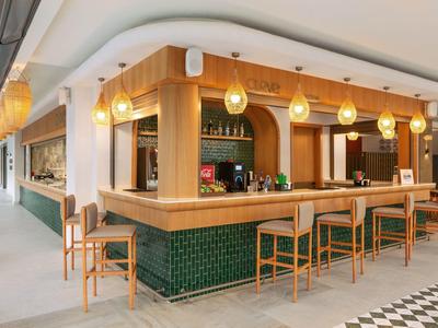 Nowoczesny bar hotelowy z drewnianym ladą, zielonymi płytkami i stołkami pod ciepłym oświetleniem.