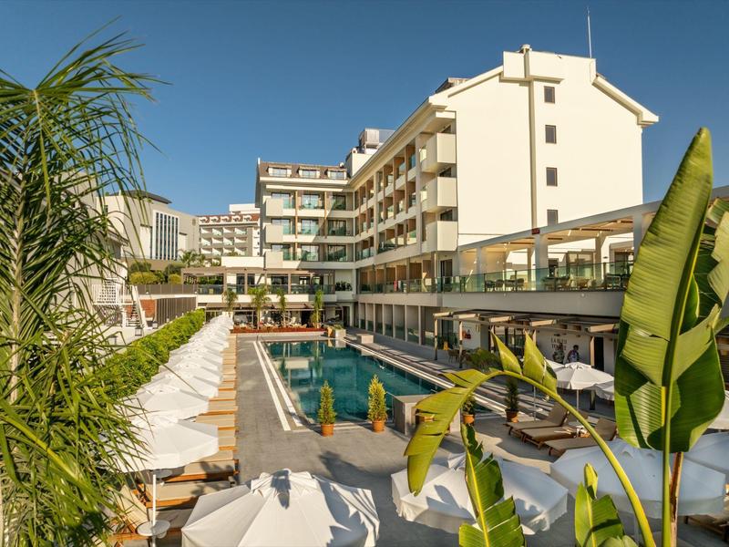 Nowoczesny kompleks hotelowy z dużym basenem, leżakami i tropikalnymi roślinami pod jasnym niebieskim niebem.